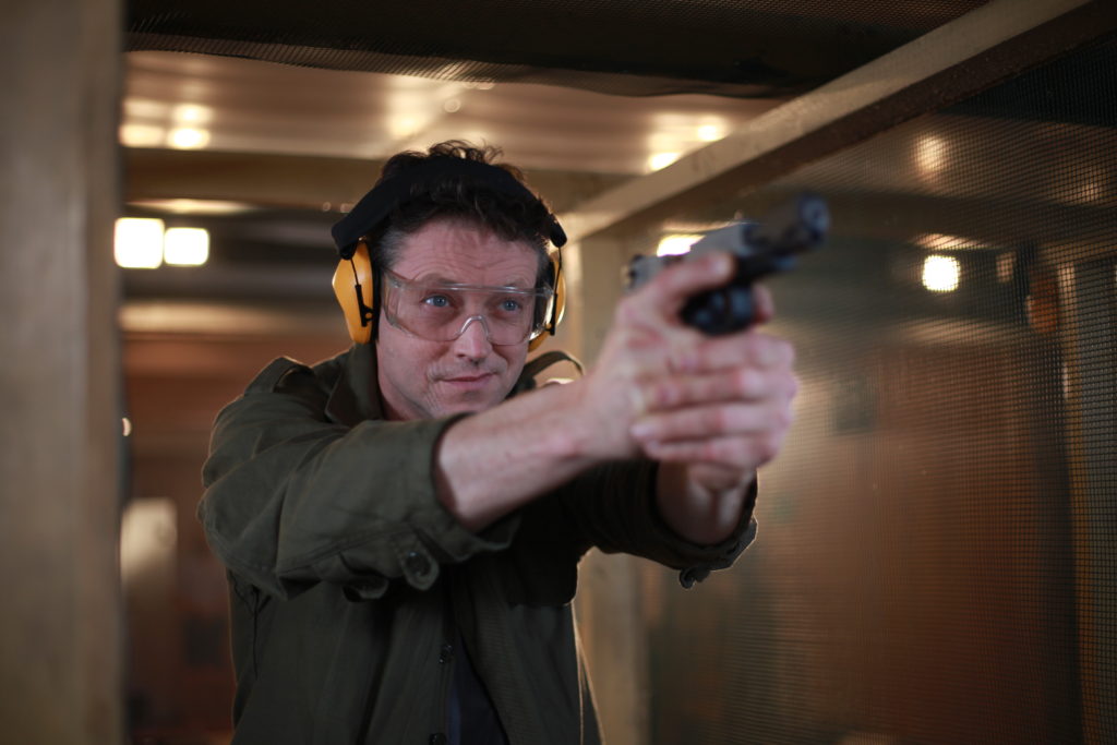 Mężczyzna w okularach, i słuchawkach ochronnych celuję z krótkiej broni na strzelnicy.