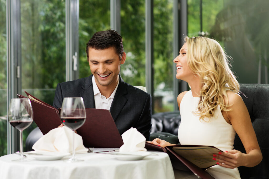Para w restauracji przygląda się karcie dań siedząc przy stoliku. Przed nimi stoją dwie lampki czerwonego wina,