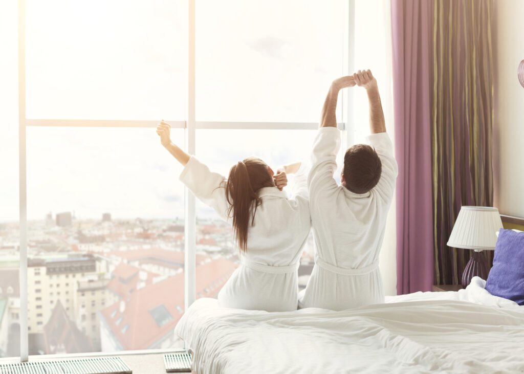 Przeciągająca się para na hotelowym łóżku w białych szlafrokach. Patrząca przez okno na widok na miasto.