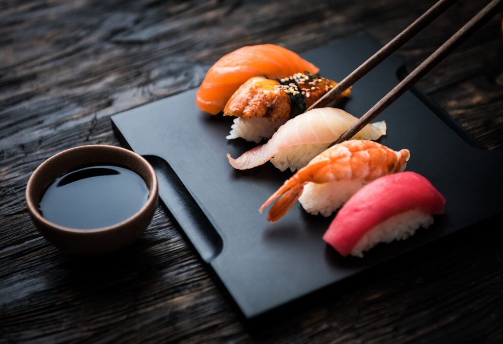 Wyeksponowane sushi na ciemnej desce z miseczką sosu sojowego. Po jeden kawałek sięgają dwie pałeczki.