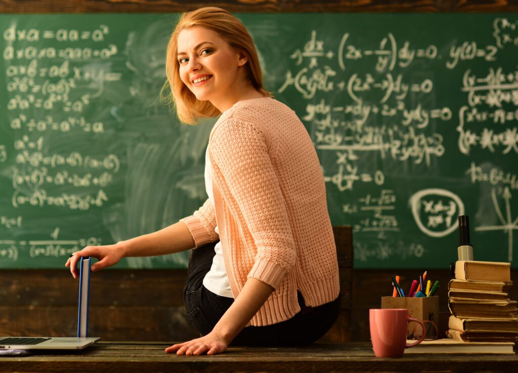 Dzień Nauczyciela - nauczycielka, w tle zapisana tablica, laptop, kubek kawy, dziennik. Prezent z okazji Dnia Nauczyciela