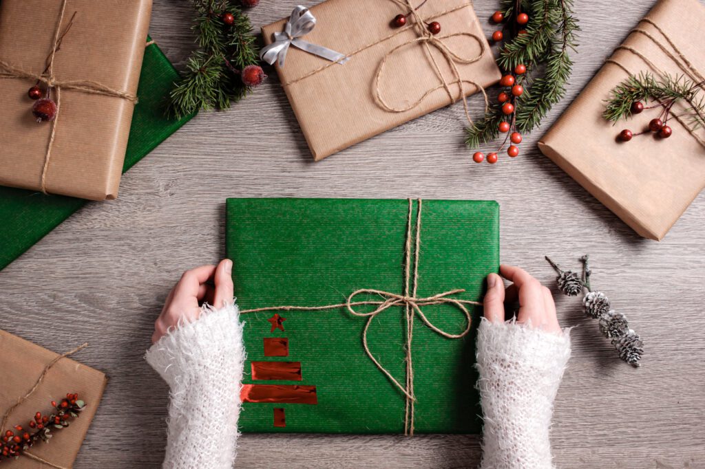 Upominki na Święta dla dalszej rodziny - dłonie kobiety w białym swetrze trzymają prezent opakowany w zielony papier