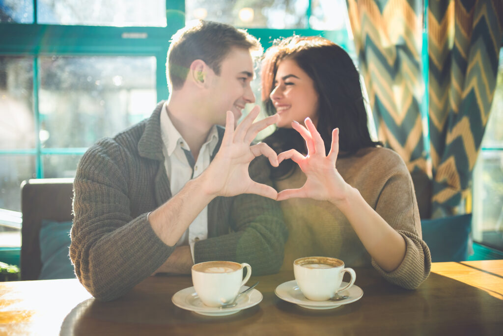 Prezent na Walentynki dla dwojga - para siedzi w kawiarni, dłońmi pokazują wspólnie serce. Na stole dwie filiżanki kawy.