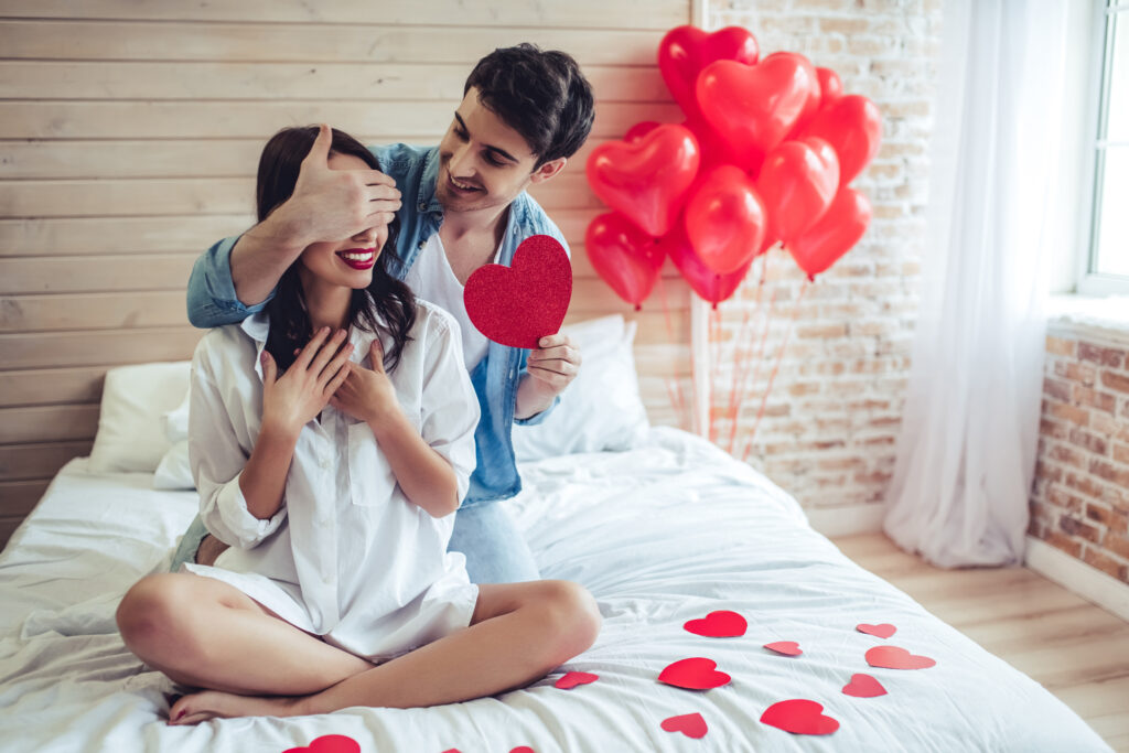 Prezent na Walentynki dla dziewczyny: Dziewczyna siedzi na łóżku po turecku, za nią klęczy chłopak, który jedną ręką zasłania jej oczy, a drugą wręcza walentynkę w kształcie serca. W tle czerwone balony w kształcie serc.