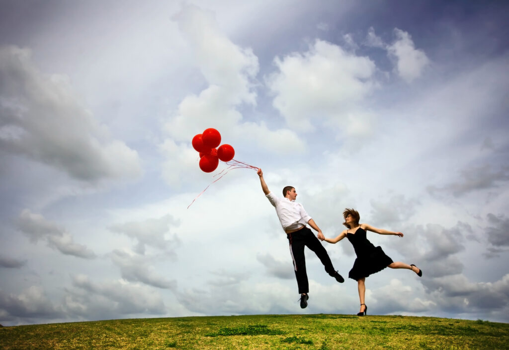 Walentynki 2023 - mężczyzna, trzyma jedną ręką czerwone balony, które go unoszą. Drugą ręką trzyma za rękę kobietę, która również zaczyna się unosić.