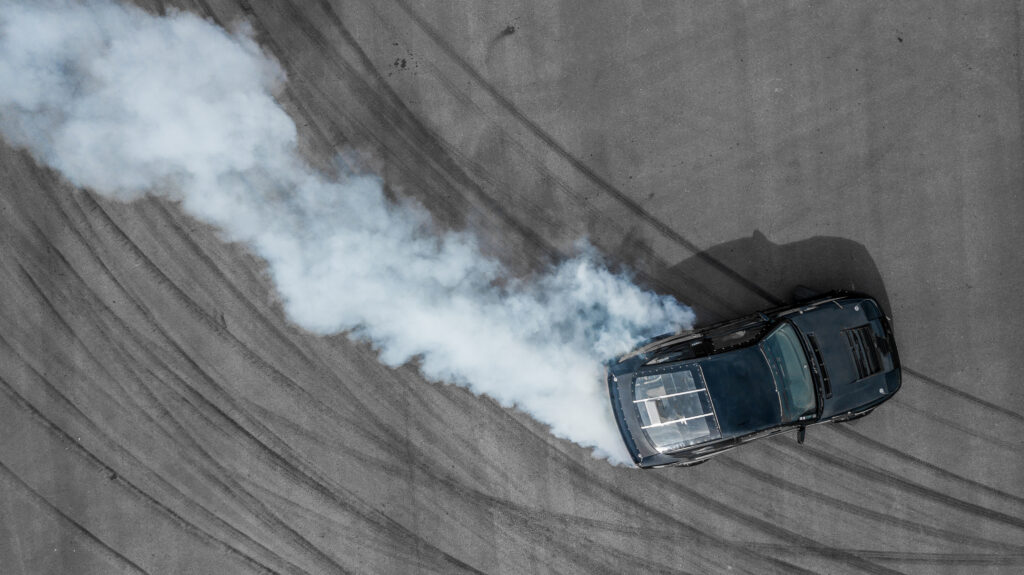 Drift: czarne auto driftuje bokiem na asfaltowym torze, zostawiając za sobą chmurę dymu. Widok z góry.
