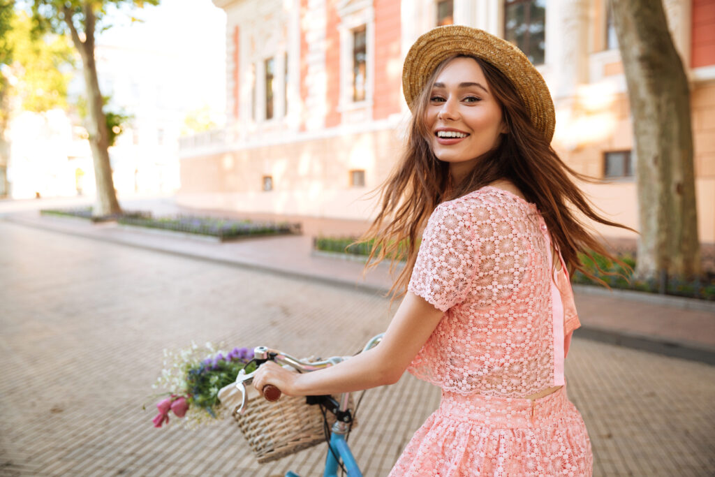 Dzień Kobiet: Kobieta jedzie na rowerze i uśmiecha się. Ma różową sukienkę i kwiaty w koszyku.