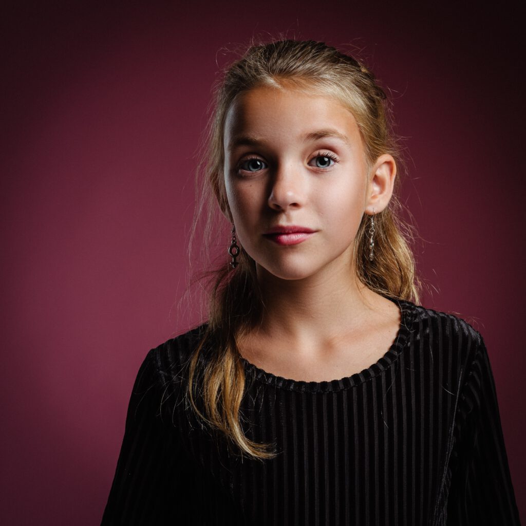 Prezent na Dzień Dziecka dla 13 latka - na zdjęciu 13-letnia dziewczynka o blond włosach, w czarnych swetrze na różowym tle. Zdjęcie portretowe.