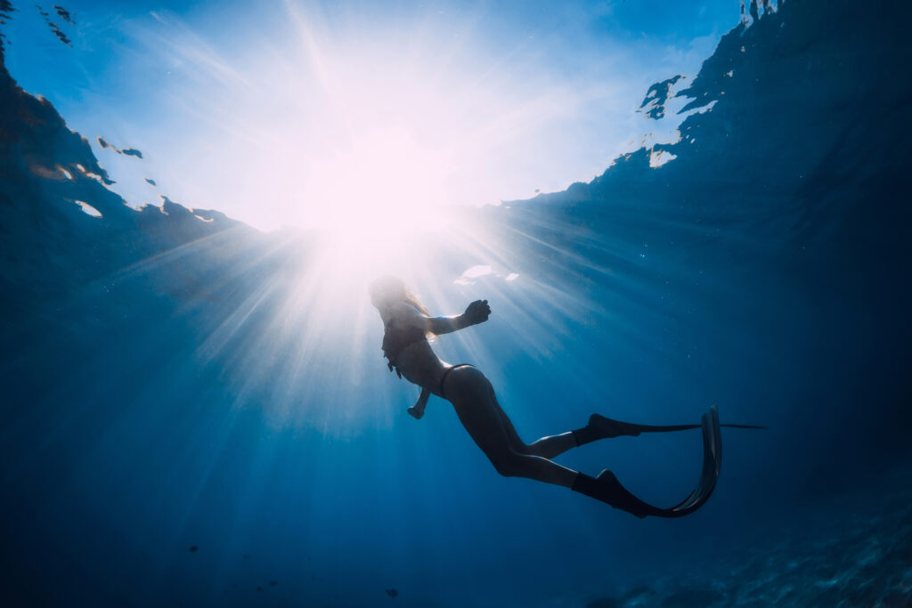 Freediving - pani w oceanie z płetwami na nogach. Wyraźnie świecące słońce przebija się w głębiny wody i zasłania twarz nurkującej kobiety.