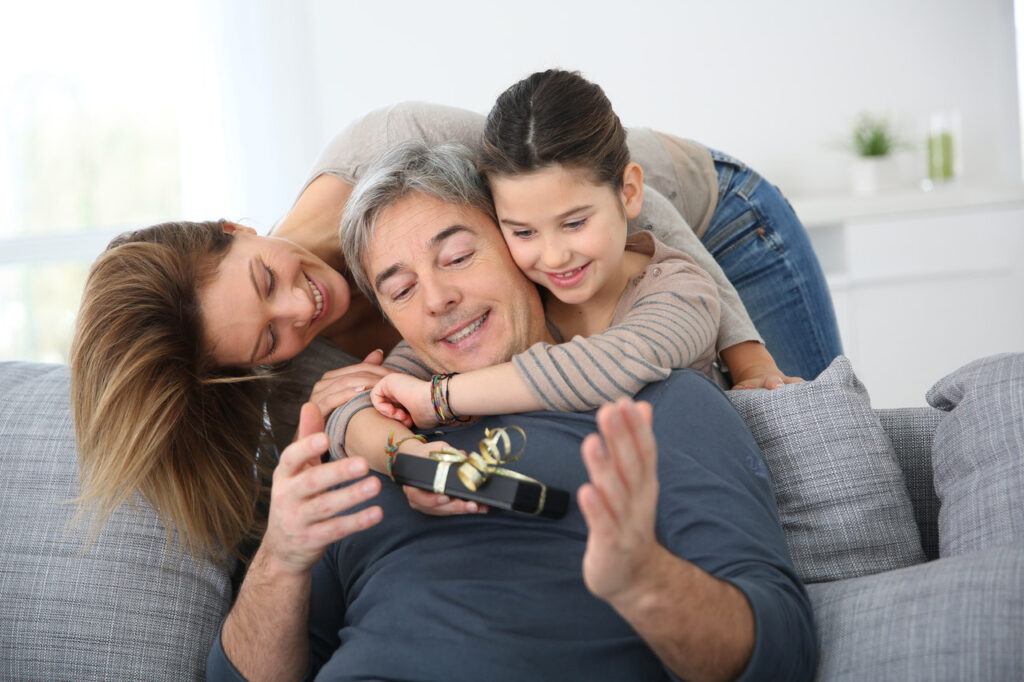 Siedzący na kanapie mężczyzna otrzymuje prezent na szczęście od przytulającej go żony i córki