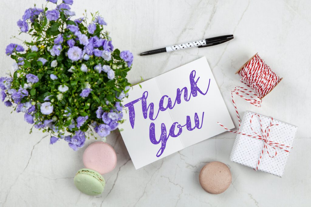 Kartka z napisem ,,thank you" leży na stole. Obok stoją kwiaty i pudełko prezentowe i leżą ciastka i szpula sznurka.