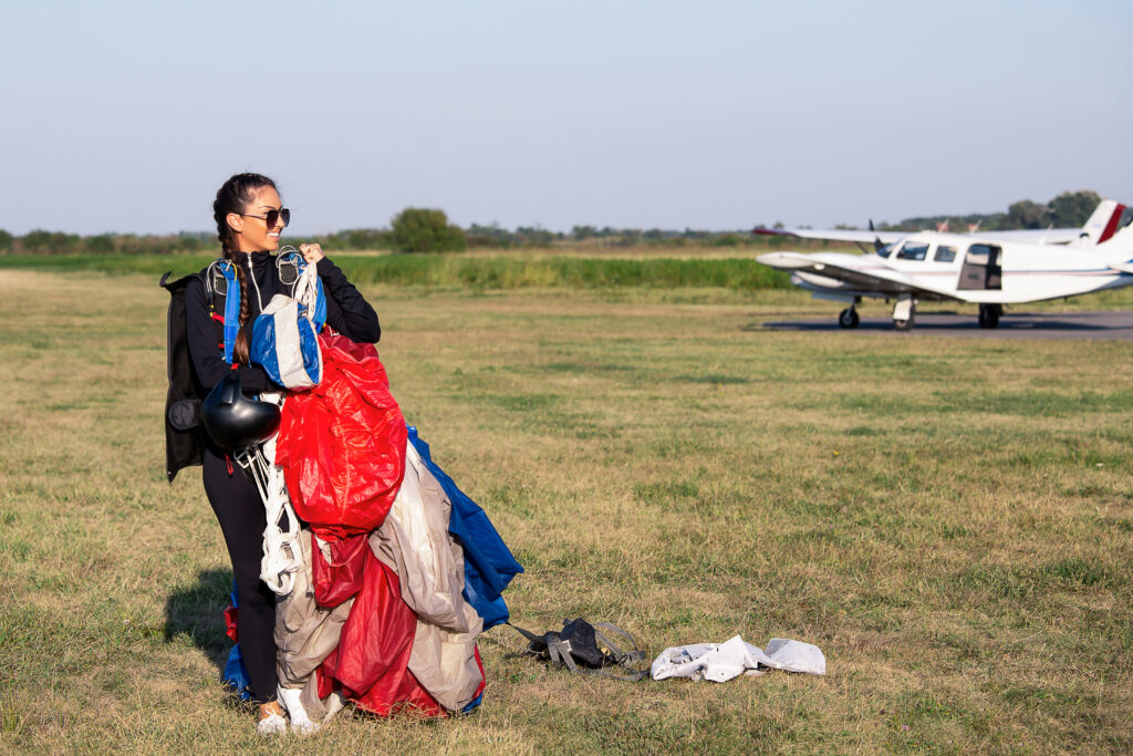 Młoda kobieta po skoku ze spadochronem. Trzyma w dłoniach sprzęt spadochronowy. W tle widać samolot.