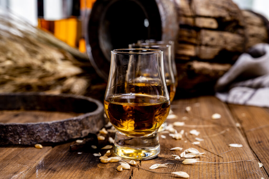 Whisky w szklankach stoi na stole. W tle drewniana beczka i snop zboża.