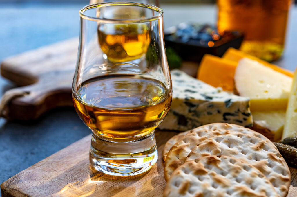 Whisky w szklance stoi na desce do krojenia. Obok leży ser, pieczywo i ogórki. W tle stół i deska do krojenia. 