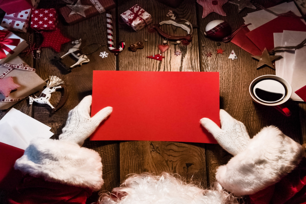 Święty Mikołaj trzyma w dłoniach czerwoną kartkę. Obok niego stoi kawa. Na stole leżą prezent, listy i ozdoby świąteczne.