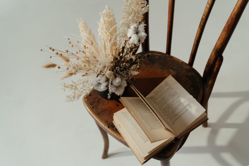 Książka leży na drewnianym krześle. Obok stoi bukiet suszonych kwiatów w wazonie. 