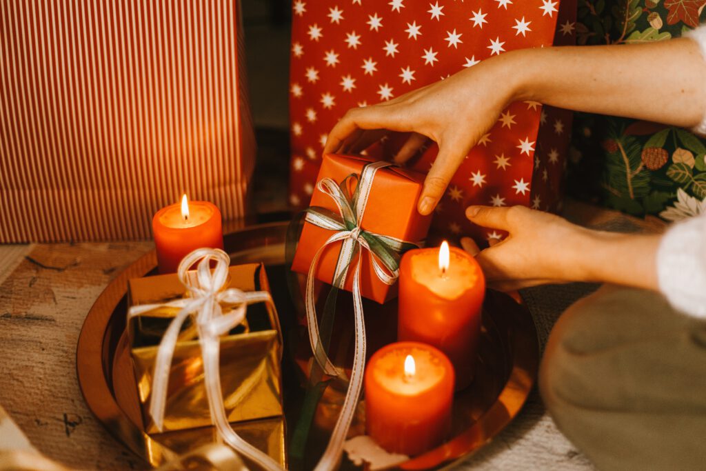 Na złotej tacy stoją zapalone świece oraz prezent. Ktoś dłonią kładzie drugi prezent na tacę. W tle inne zapakowane prezenty.