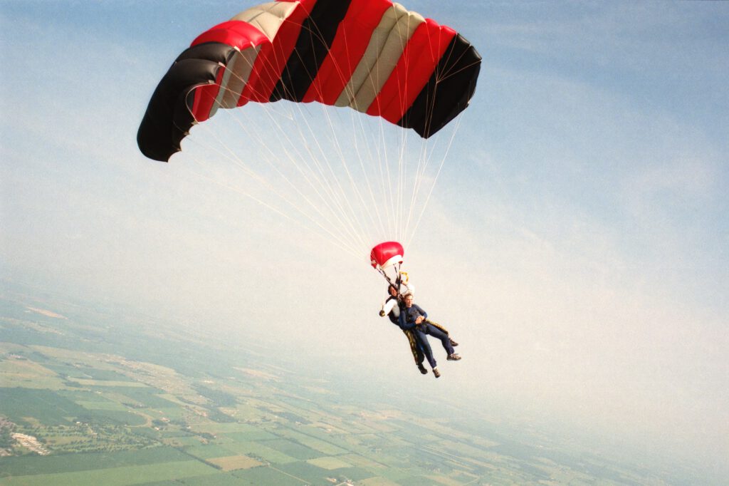 Dwie osoby skaczą ze spadochronem. W tle niebo, pola i wsie.