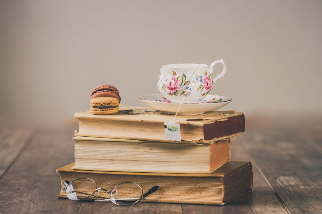 Trzy książki leżą na drewnianym stole. Przed nimi leżą okulary. Na książkach znajdują się dwa ciastka i filiżanka z herbatą.