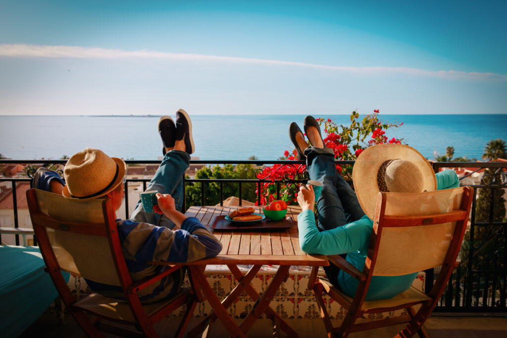 Mężczyzna i kobieta siedzą na balkonie i patrzą na morze. Siedzą na krzesłach, z nogami położonymi na barierkę, a w dłoniach trzymają kubki.