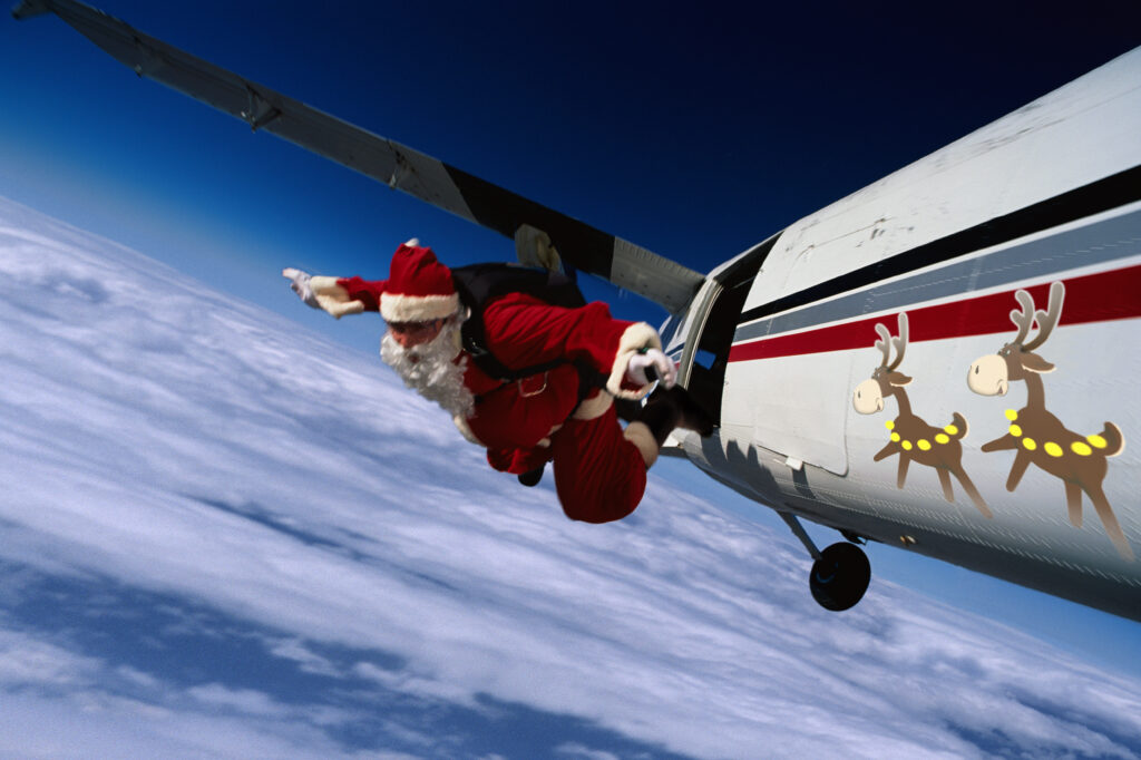 Św. Mikołaj skacze z samolotu. Samolot ozdobiony jest w rysunki reniferów. W tle niebo.