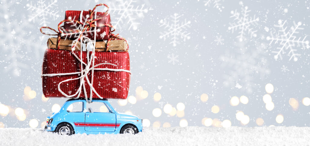 Mały niebieski samochód wiezie na dachu prezenty świąteczne. W tle pada śnieg.