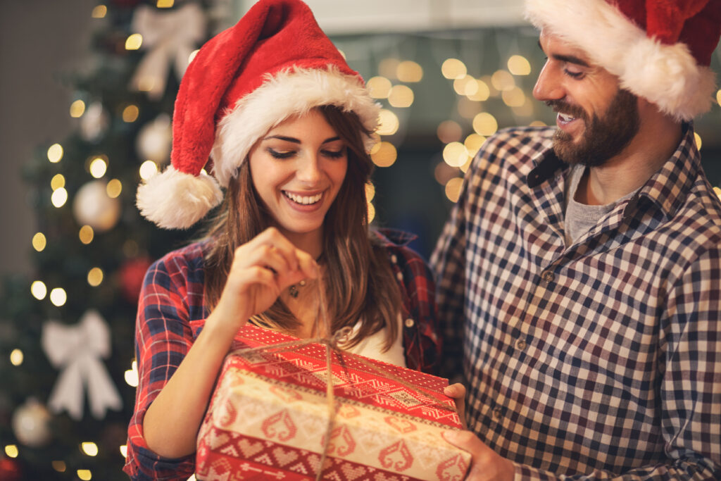 Mężczyzna i kobieta ubrani w czapki Mikołaja otwierają prezent. W tle znajduje się oświetlona choinka.