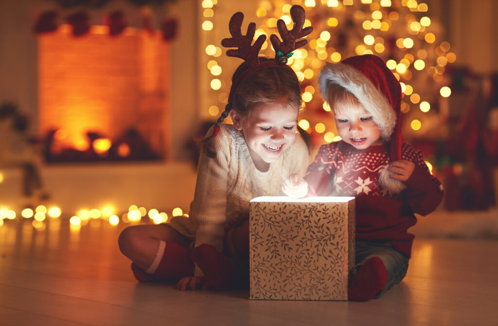 Chłopiec i dziewczynka otwierają prezent. W tle lampki choinkowe na podłodze, kominek i przystrojona choinka.