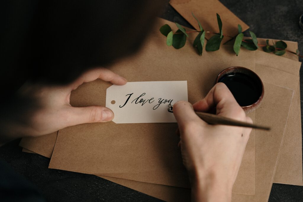 Osoba pisze piórem na karteczce ,,I love you". W tle miseczka z atramentem, brązowy papier i gałązka z listkami.