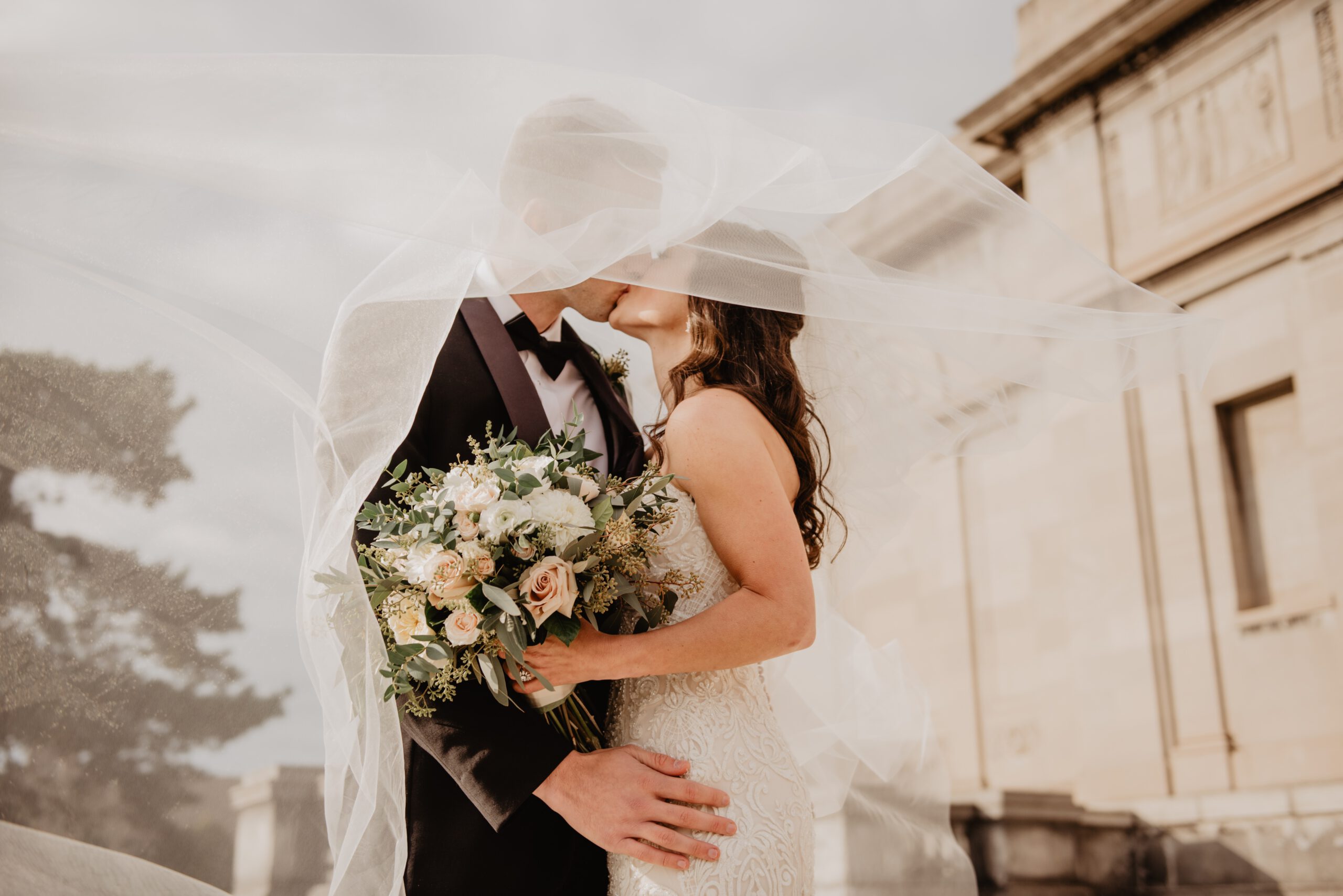 Małżeństwo całuje się i przytula.  Kobieta ma na sobie białą ślubną suknię i kwiaty w dłoniach. Mężczyzna ma garnitur. Ich twarze zakrywa rozwiany welon.