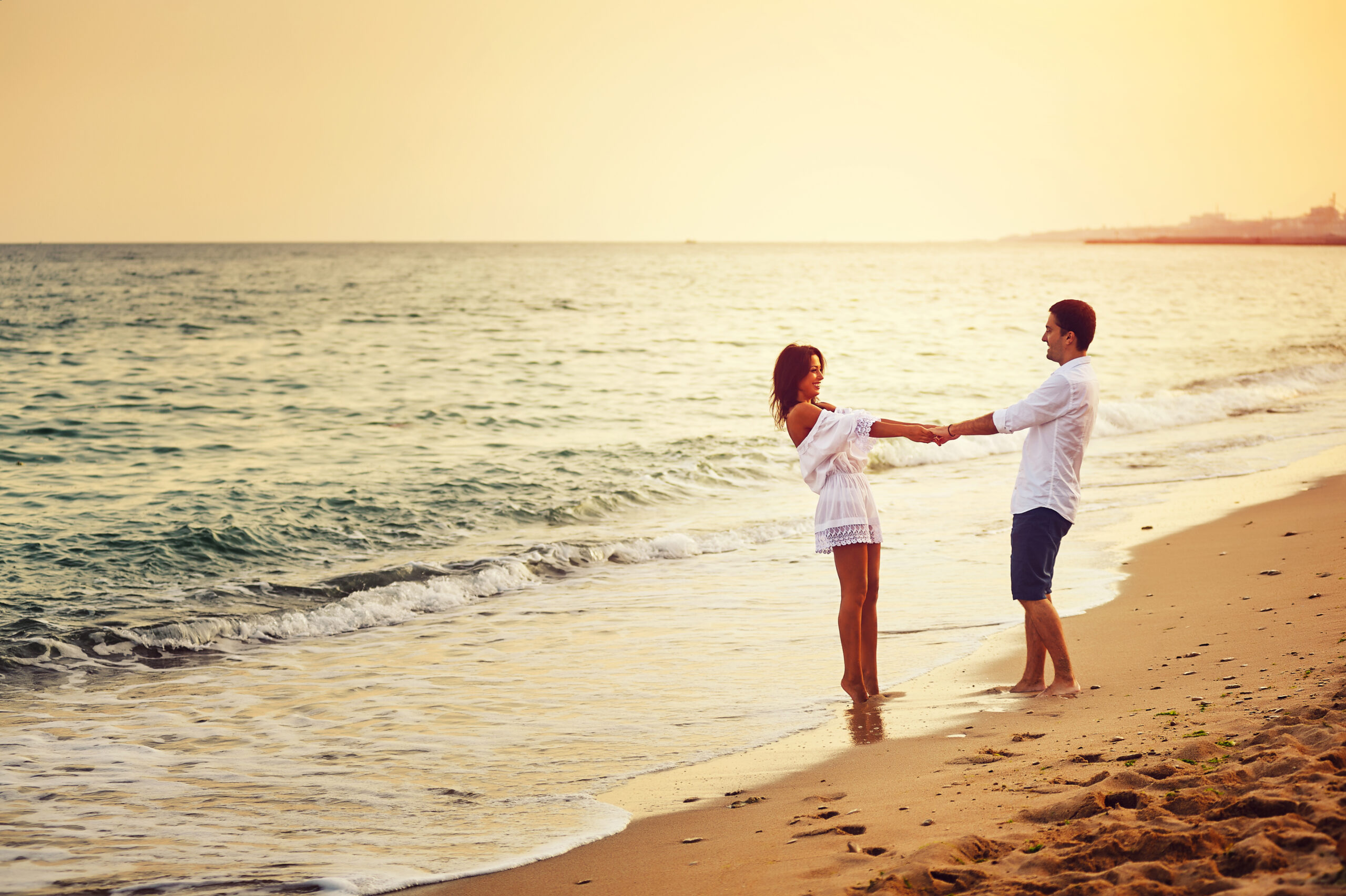 Mężczyzna i kobieta stoją na plaży i trzymają się za ręce. Patrzą na siebie. W tle morze.