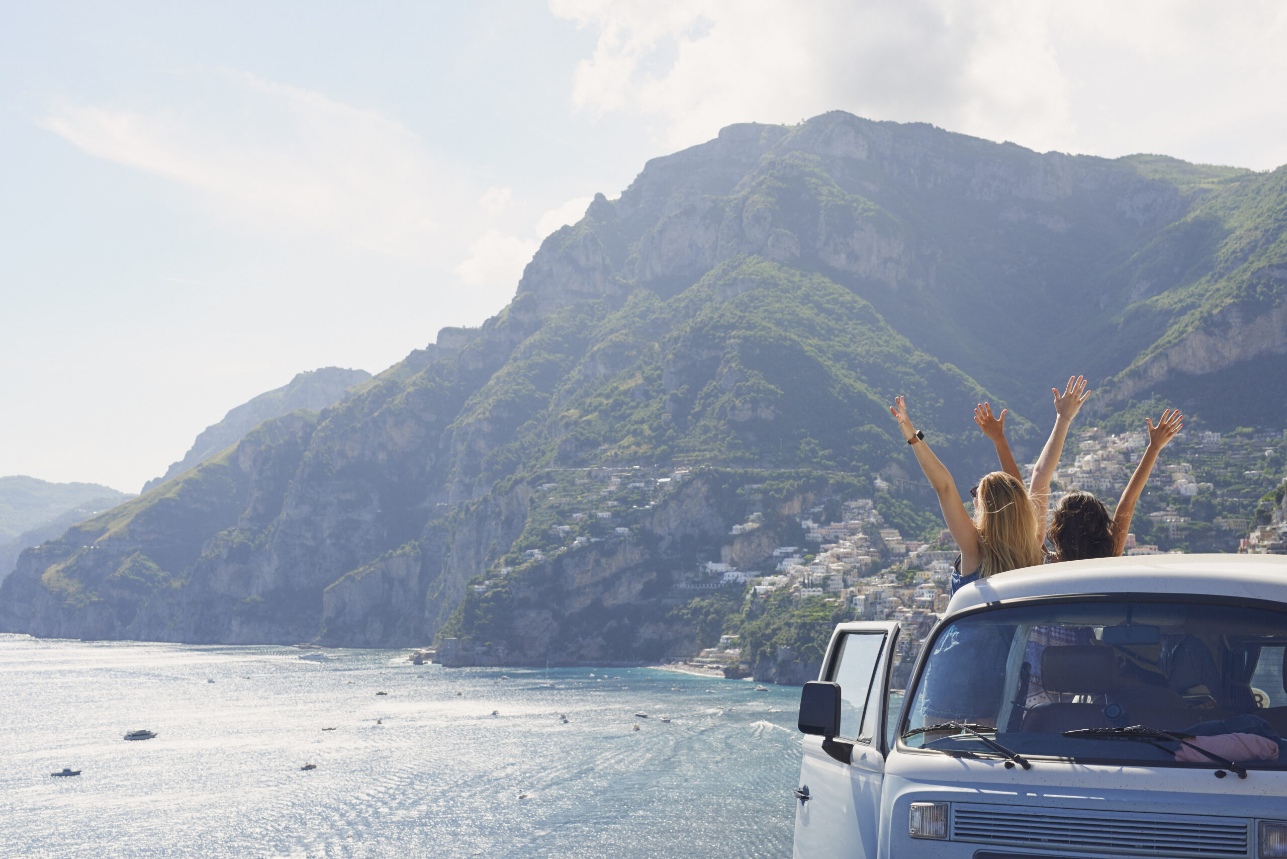 Dwie kobiety stoją przed samochodem i unoszą ręce. Przed nimi morze, góry i niewielkie miasteczko.