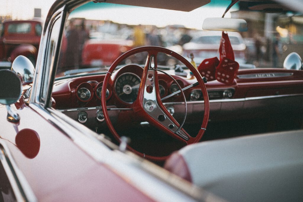 Wnętrze starego auta. Czerwona kierownica i deska rozdzielcza, na lusterku wisi breloczek z czerwonymi kostkami do gry.