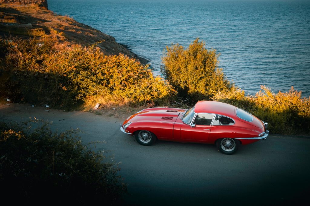 Czerwony Jaguar E-type stoi na drodze. W tle znajduje się wzgórze i morze.
