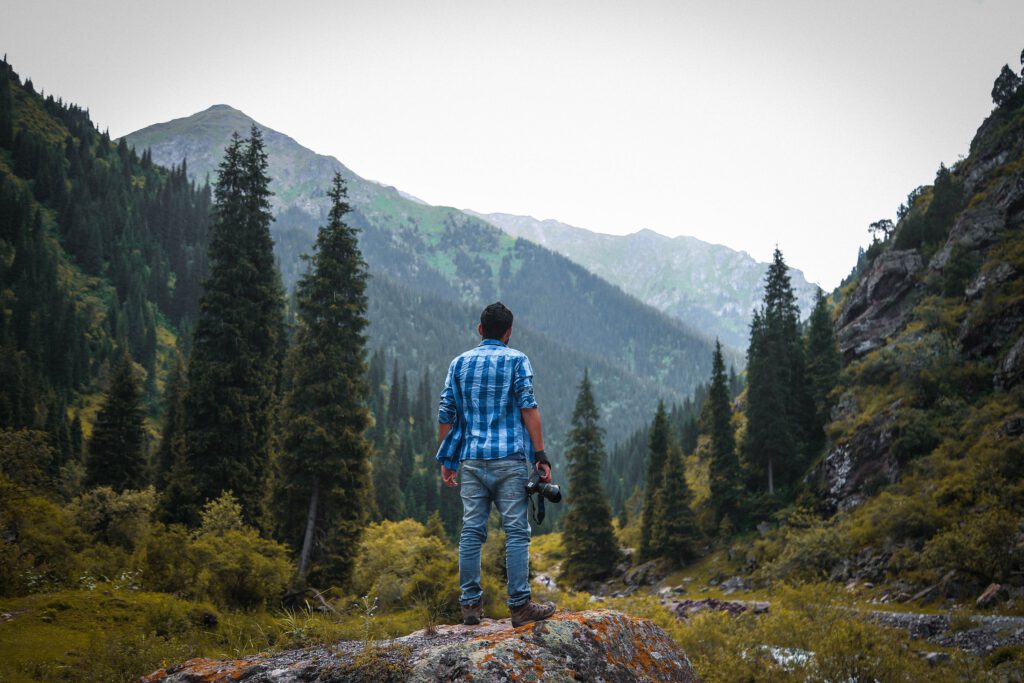 Mężczyzna stoi tyłem na skale i patrzy w dal. W prawej dłoni ma aparat fotograficzny. Ubrany jest w jeansy i niebieską koszulę. W tle góry, lasy, trawy i doliny.