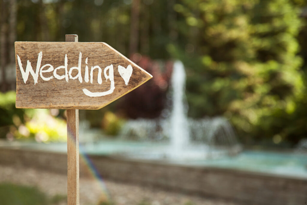 Drewniana tabliczka z napisem ,,Wedding" i serduszkiem. W tle fontanna i drzewa.