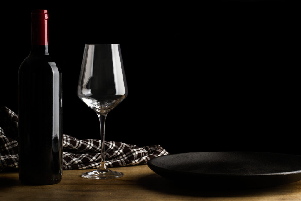 Na drewnianym stole stoi czarny talerz. Po lewej stronie znajduje się pusty kieliszek, czarna butelka od wina, a za nimi leży czarno-biała szmatka w kratkę. Za nimi znajduje się czarne tło.