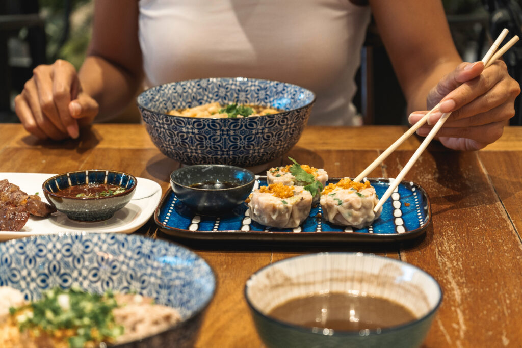 Kobieta je pałeczkami sushi, siedząc przy stole. Na nim stoją też miski z zupą i sosami.