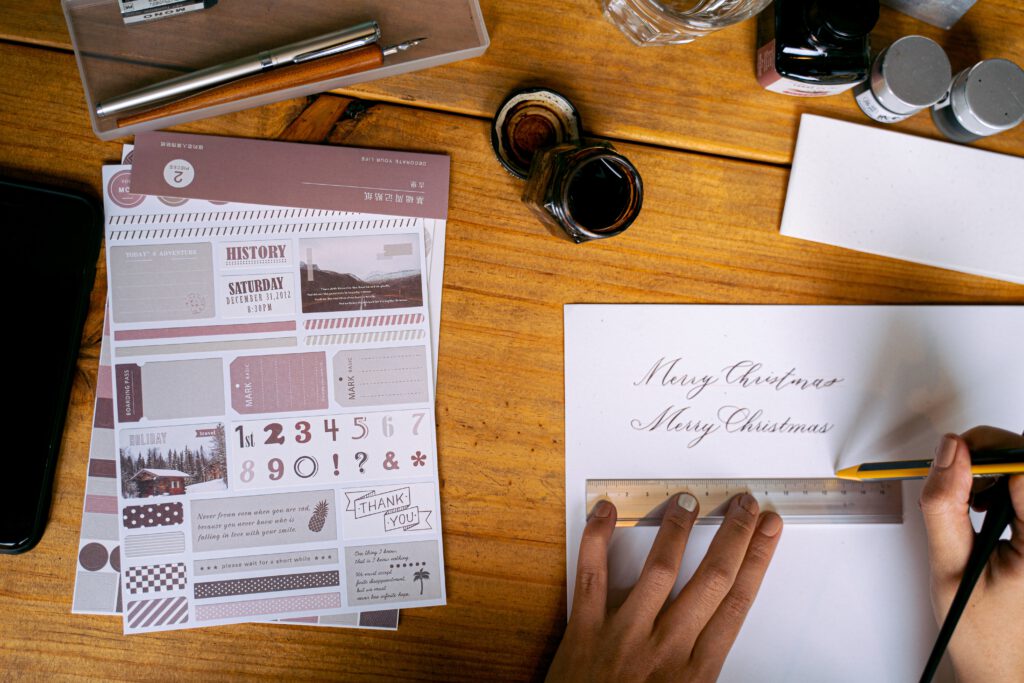 Na biurku leżą butelki atramentu, kartka papieru, naklejki i pióro. Ktoś pisze na kartce ,,Merry Christmas" 
