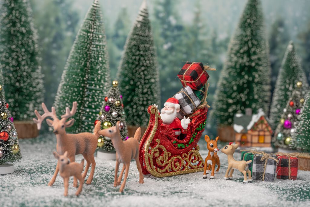 Figurki Świętego Mikołaja w saniach z prezentami i reniferów. W tle figurki ośnieżonych drzew i domku.