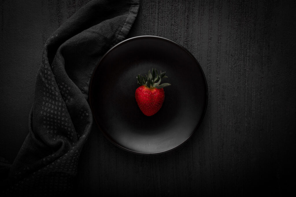 Na czarnym stole leży czarny talerz a obok czarny materiał. Na talerzu znajduje się czerwona truskawka. 