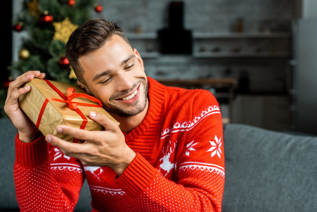 Mężczyzna w czerwonym, świątecznym swetrze siedzi na kanapie. Trzyma w rękach świąteczny prezent i przykłada go do ucha. W tle widać kuchnię i choinkę.
