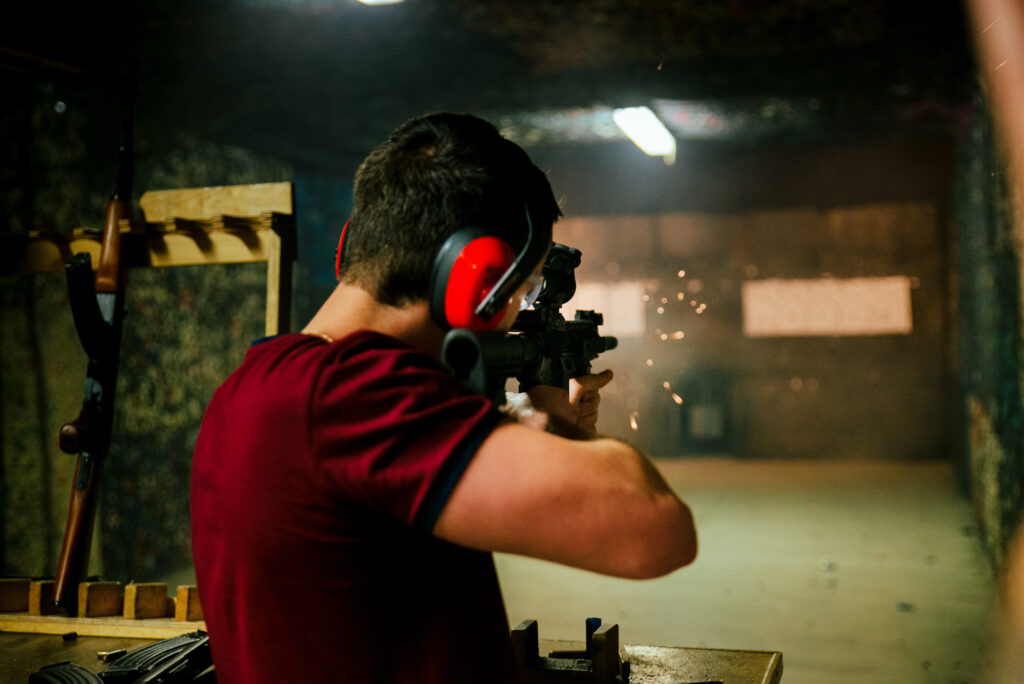 Mężczyzna jest na strzelnicy. Strzela z broni. Ubrany z czerwoną koszulkę, a na uszach ma słuchawki do strzelania. Po lewej widać drewniany stojak, w którym stoi broń.