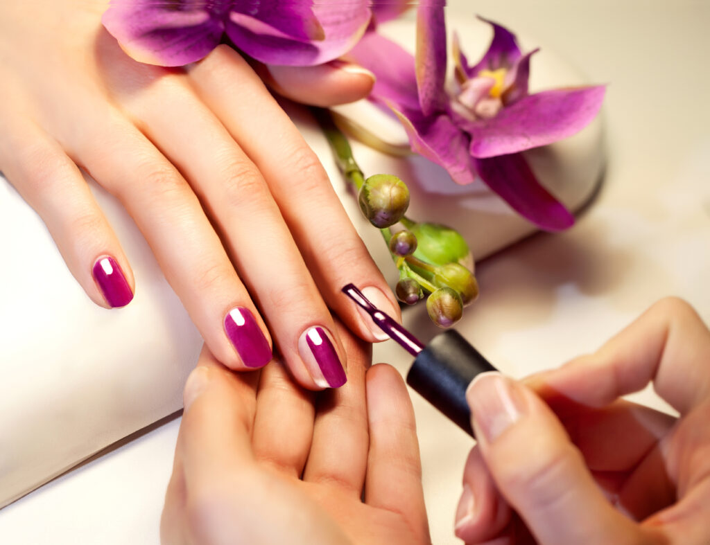 Kobieta maluje drugiej kobiecie paznokcie na kolor fioletowy. Dłonie spoczywają na białej poduszce. Obok leży fioletowy kwiat.