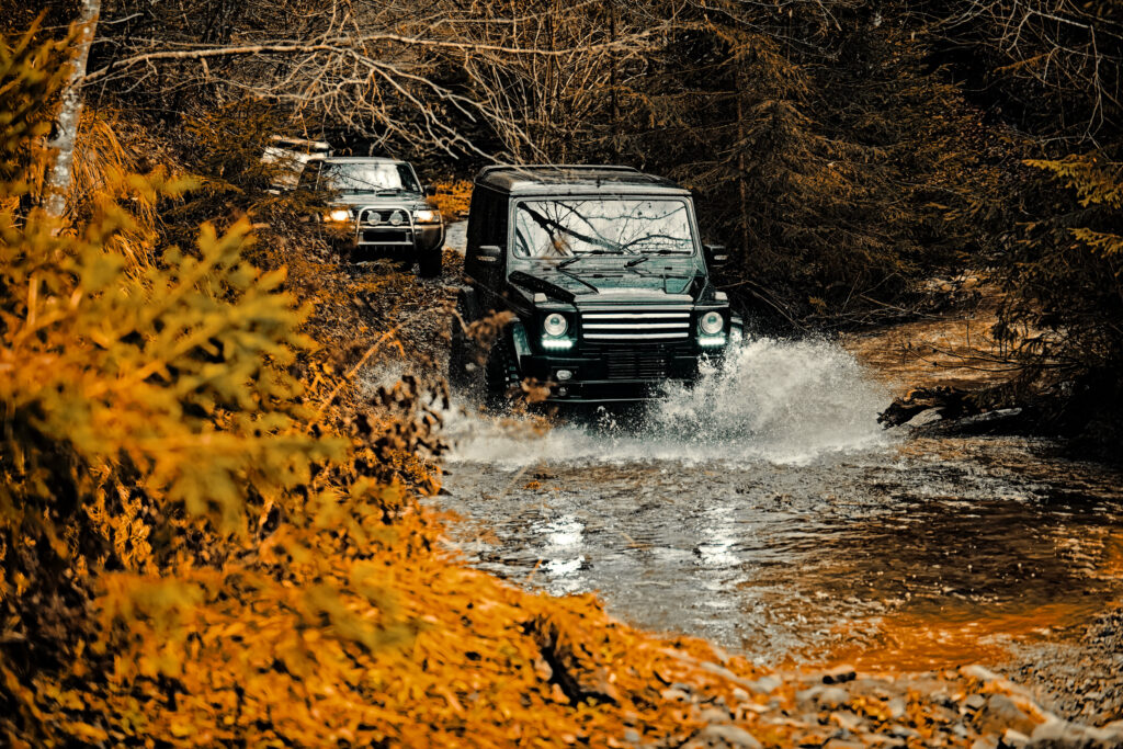 Dwa samochody terenowe jadą jeden za drugim po ścieżce leśnej zalanej wodą. Dookoła jesienny las.