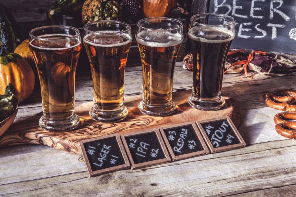 4 szklanki z piwem stoją na drewnianej podstawce. Przed nimi leżą tabliczki z napisanymi nazwami piw. Dookoła leżą precle, kukurydza i dynie.