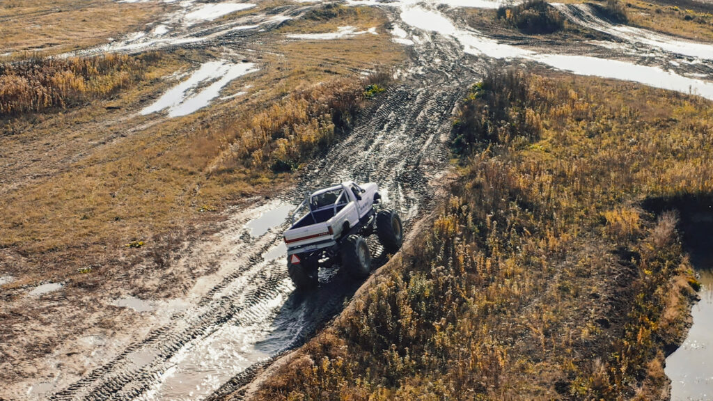 Biały Monster Truck pokonuje wzniesienie na błotnistym torze - widok z góry, nagrany dronem, na tył pojazdu