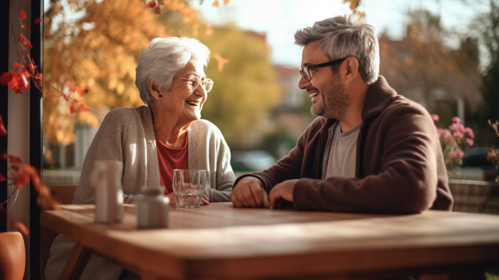 Starsza kobieta w okularach i mężczyzna w okularach siedzą przy stole. Śmieją się. Na stole stoi szklanka. W tle okno, drzewa i kwiaty.
